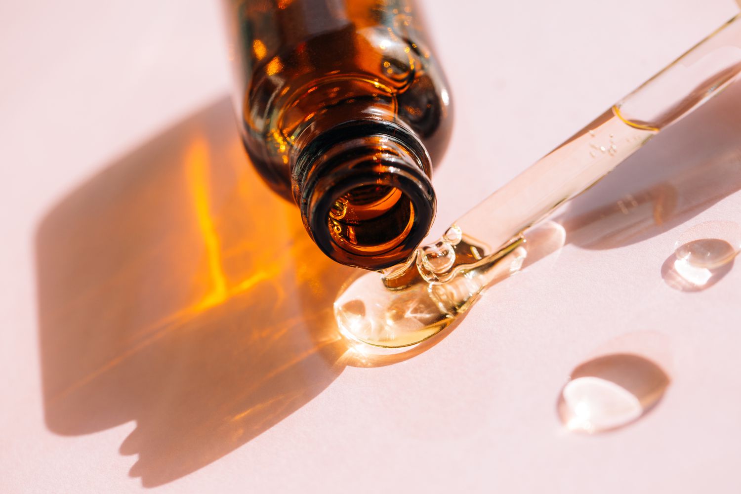 Drops of facial serum or other skincare product pouring out of an amber glass bottle, on pink background. 