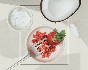 A mashed strawberry in a bowl alongside an opened coconut and coconut flakes on an geometric designed background 